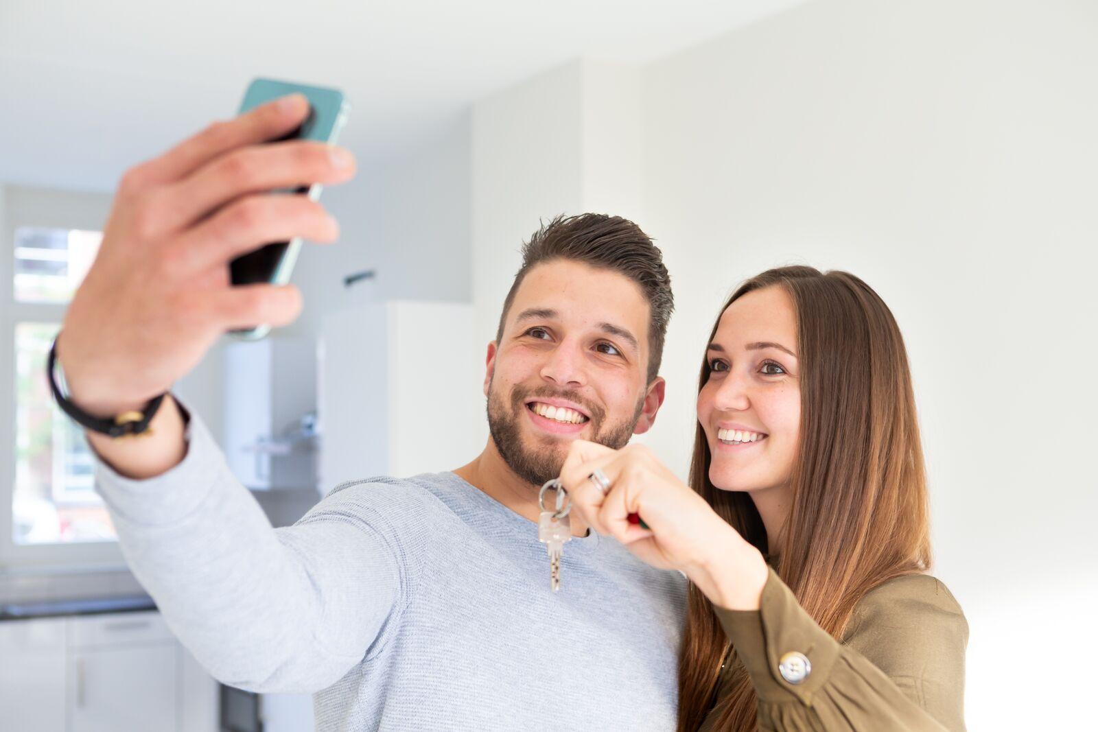 een man en een vrouw die een selfie maken met sleutels van hun nieuwe woning in de hand.
