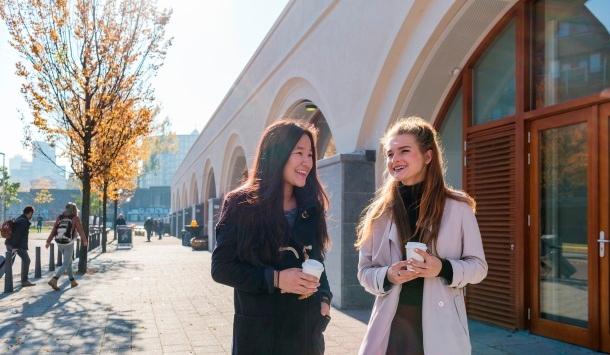Twee vrouwen lopen lachend met een kopje koffie op straat.