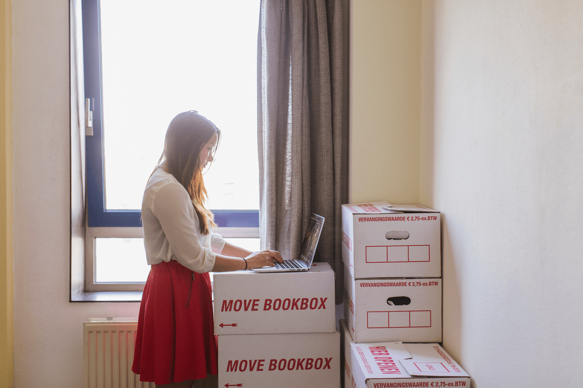 Een vrouw naast opgestapelde verhuisdozen met een laptop op de dozen. Ze zoekt naar een nieuwe woning. 