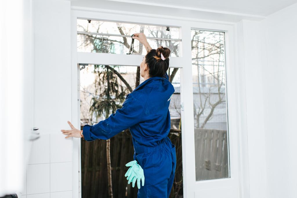 Een vrouw in een blauwe overal en schoonmaakhandschoenen maakt een raam open.